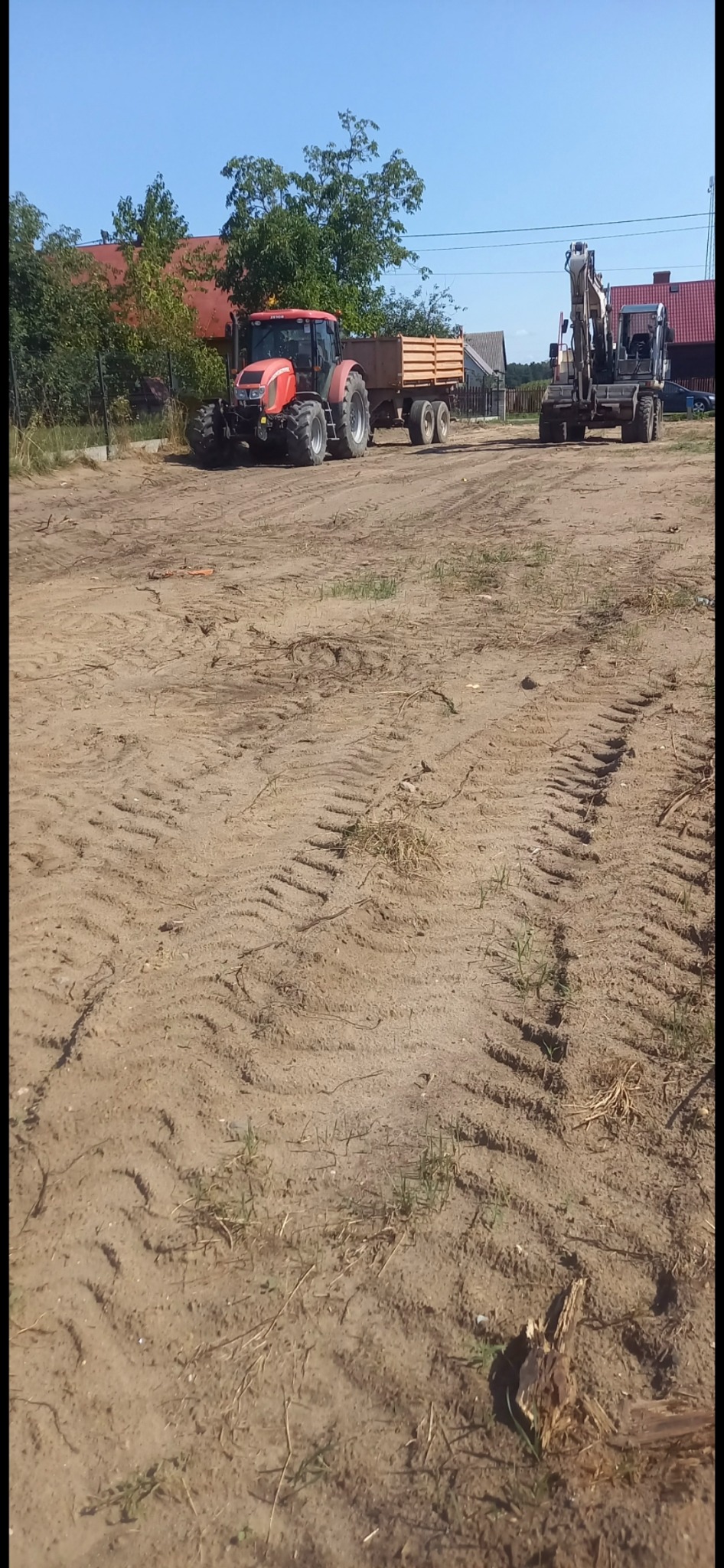 Czerwony traktor z przyczepą i koparka na placu budowy. Ślady opon na ziemi, w tle zabudowania i drzewa w słoneczny dzień. Prace ziemne w toku.