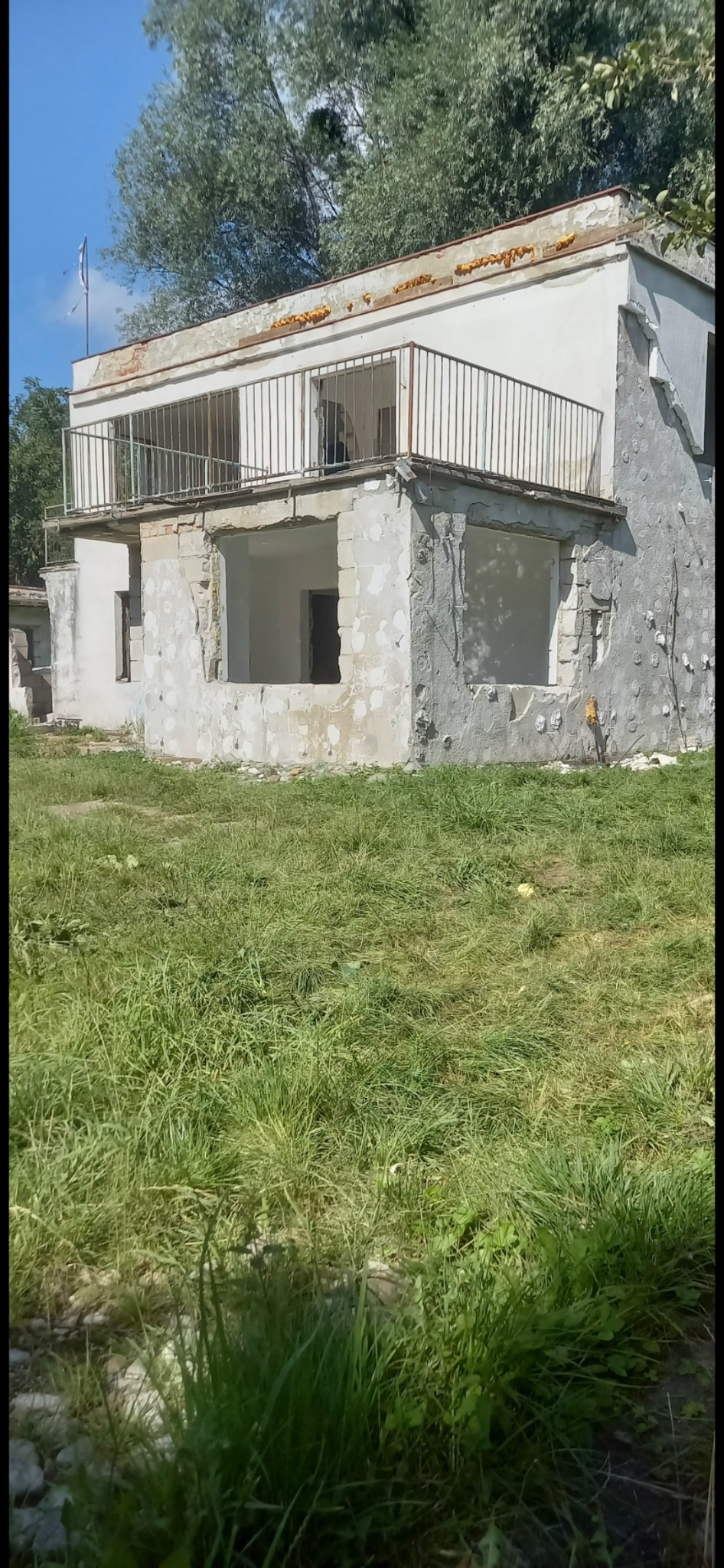 Dwukondygnacyjny budynek w trakcie rozbiórki, z widocznymi otworami okiennymi bez okien i balkonem z metalową barierką, otoczony wysoką trawą.