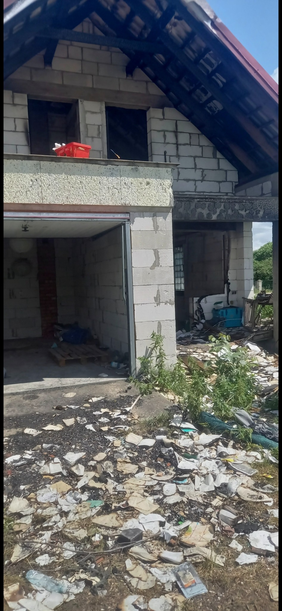 Niedokończony budynek z pustaków, z otwartym garażem i gruzem na podwórku. Czerwony koszyk na dachu garażu. Widoczne ślady po pożarze na elementach konstrukcyjnych.