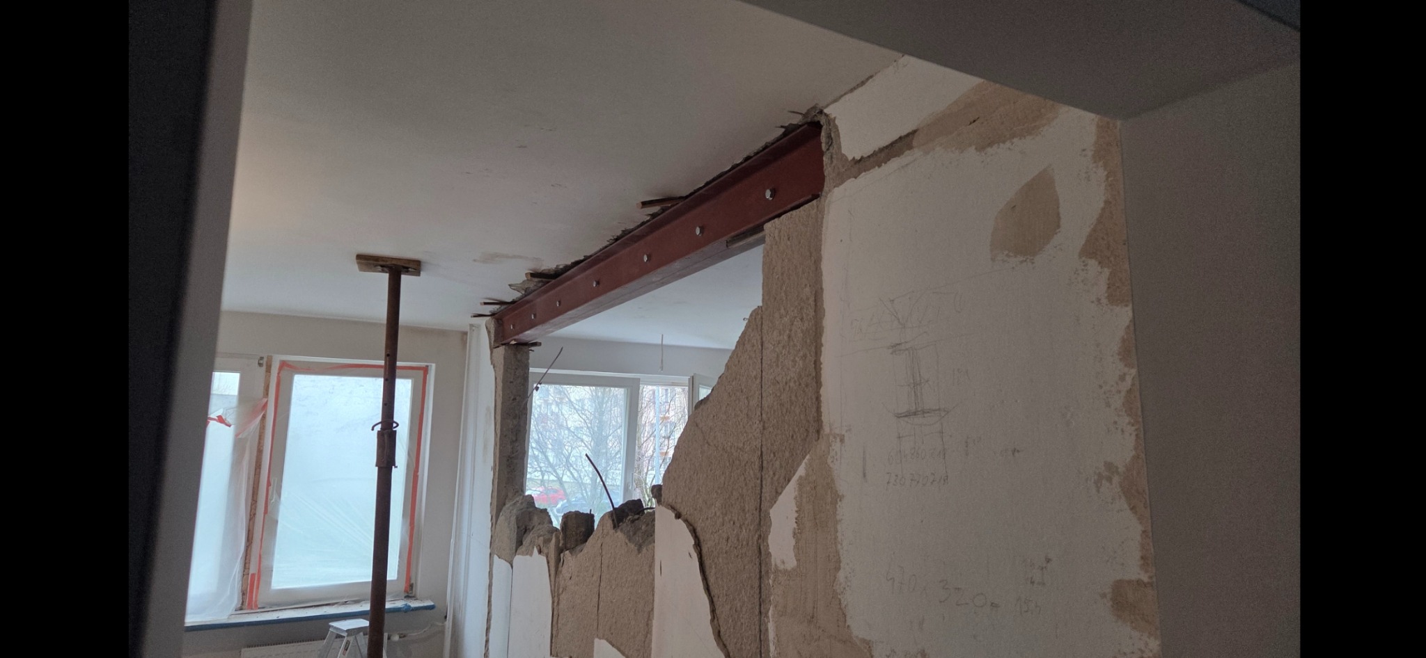 Wyburzona ściana z widoczną stalową belką nośną, podpartą stemplem budowlanym. Na ścianie widoczne odręczne notatki. Okno zabezpieczone folią malarską.