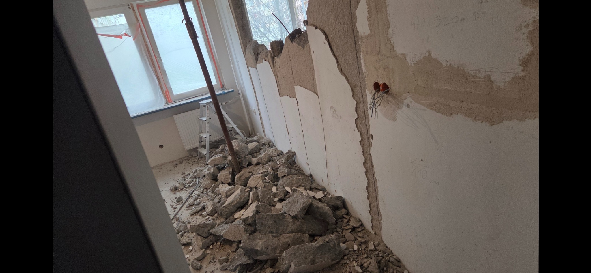 Wnętrze w trakcie wyburzania ściany: gruz na podłodze, odsłonięte kable elektryczne, częściowo zdemontowana ściana z widocznymi fragmentami instalacji.
