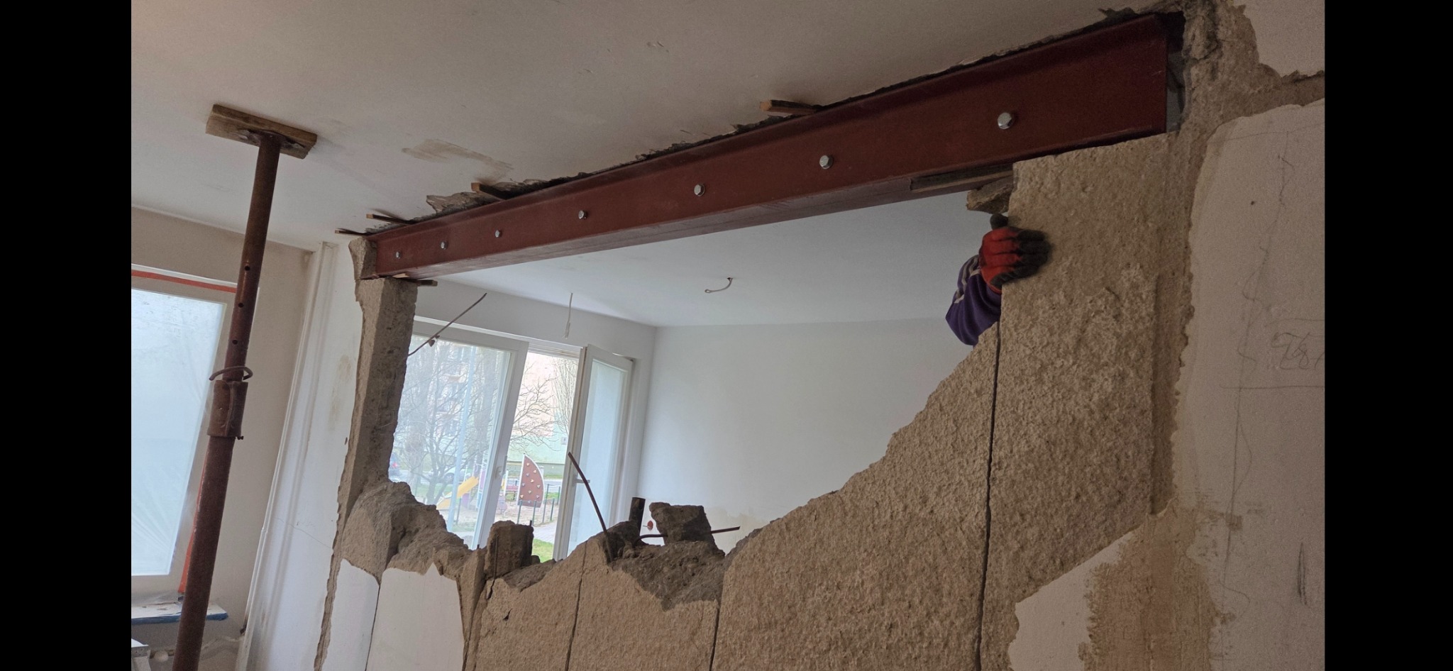 Wyburzona ściana z widoczną stalową belką wzmacniającą, podpartą stemplem budowlanym. Fragment ściany z gruzem, okno w tle. Prace wyburzeniowe w trakcie.