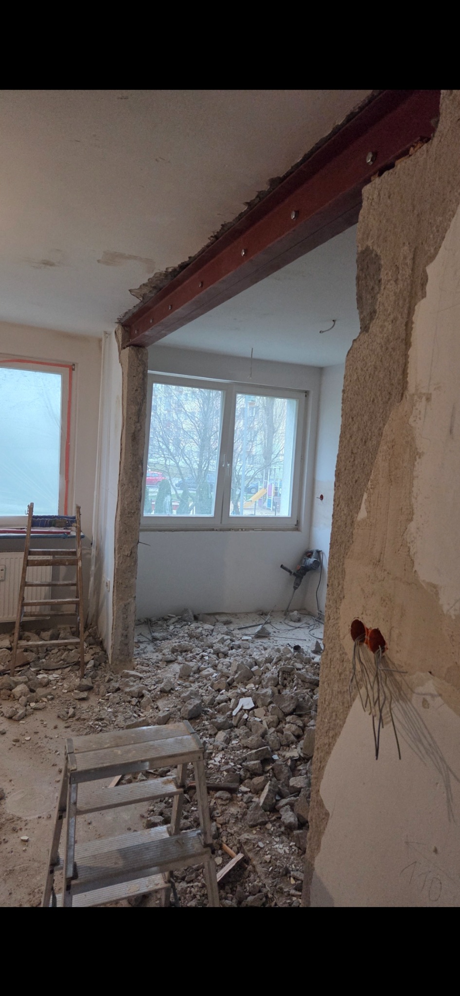 Wnętrze w trakcie wyburzania ściany, z widoczną stalową belką wzmacniającą, gruzem na podłodze i narzędziami. Widok na okno z zewnątrz.