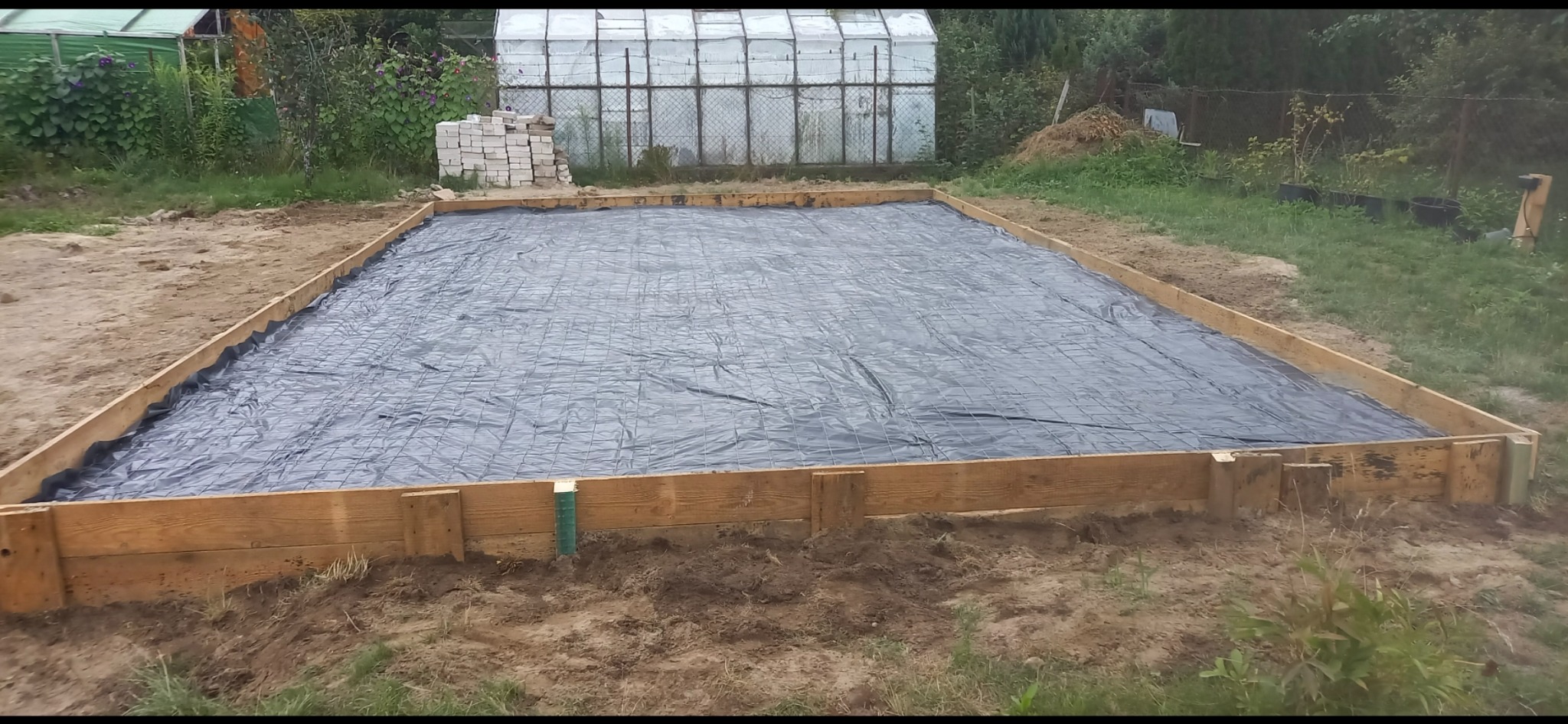 Przygotowany teren pod wylewkę fundamentów: szalunek z desek, folia i siatka zbrojeniowa. W tle szklarnia i cegły.