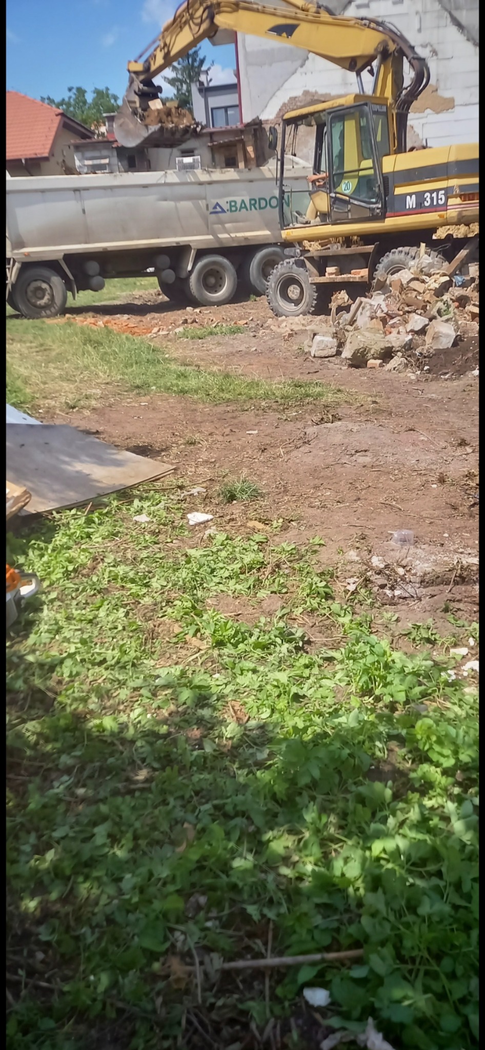 Żółta koparka CAT M315 ładuje gruz do białej ciężarówki A.BARDON, w tle fragment budynku w trakcie rozbiórki, niebo z lekkimi chmurami.