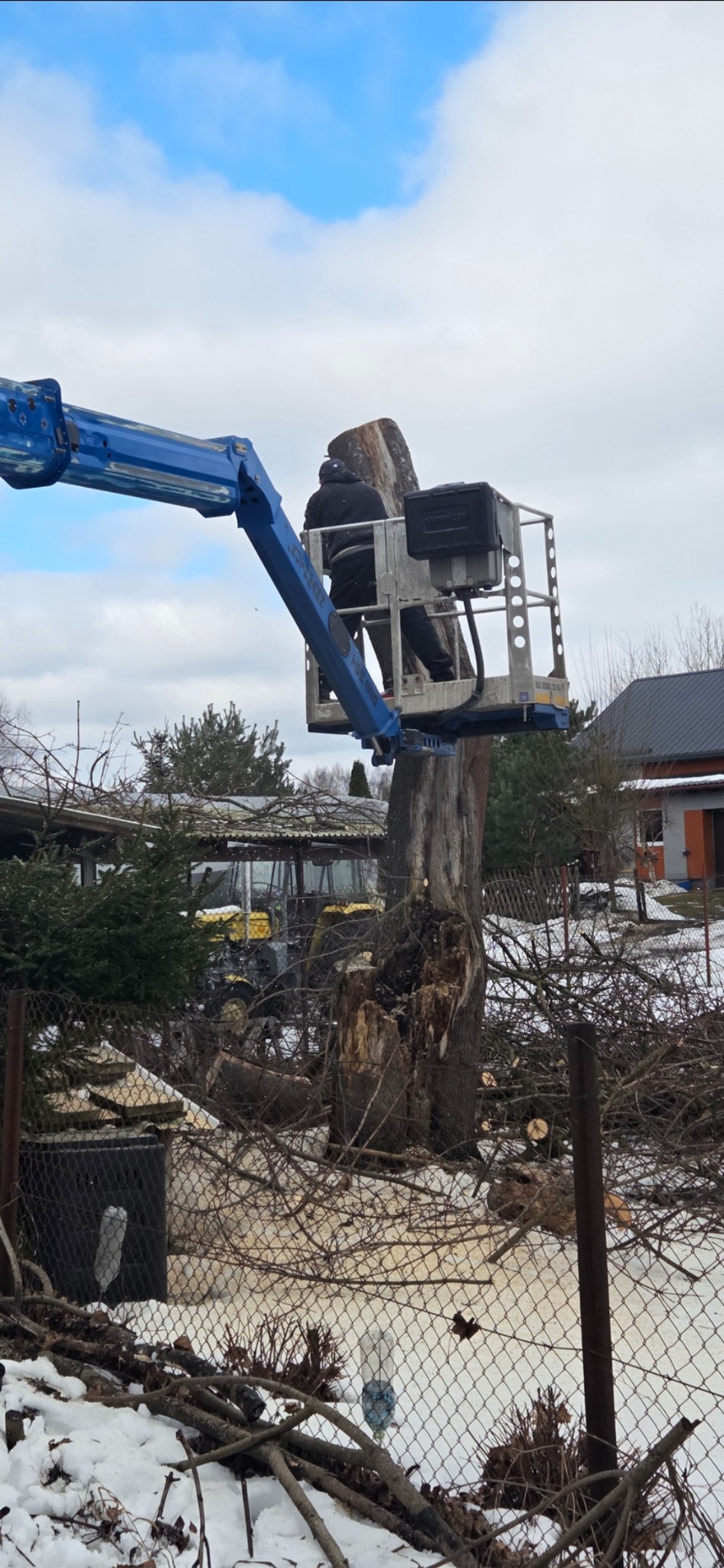 Pracownik na podnośniku koszowym tnie resztki pnia drzewa. W tle widoczny traktor i budynki. Wokół pnia leżą gałęzie i śnieg.
