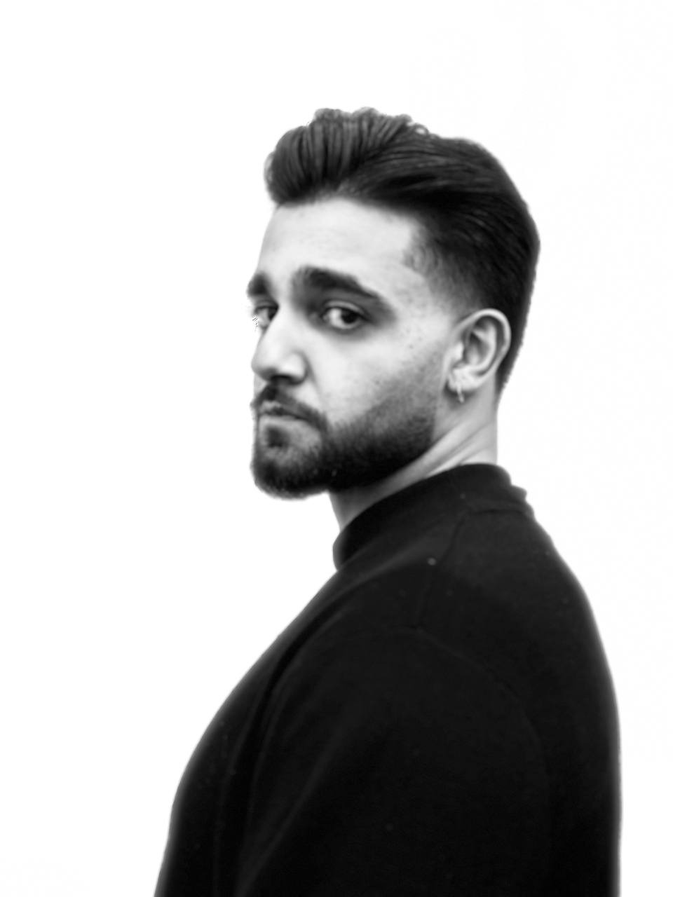 Mężczyzna z modną fryzurą, krótko ostrzyżonymi bokami i dłuższą górą, spogląda przez ramię na czarno-białym portrecie.