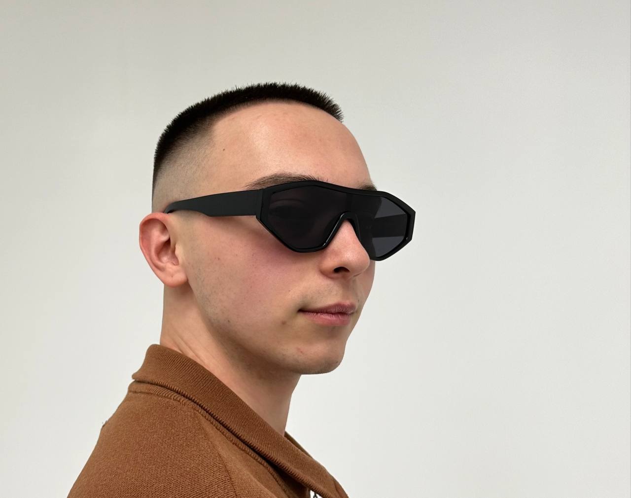 Mężczyzna z krótkimi włosami i geometrycznymi okularami przeciwsłonecznymi w brązowej koszulce polo na tle białej ściany.