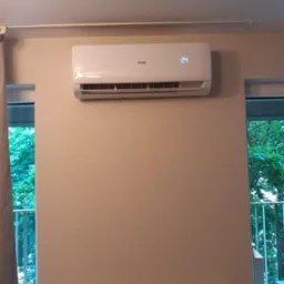 Biała klimatyzacja Haier zamontowana na ścianie w pomieszczeniu, wyświetlająca temperaturę 24 stopnie, widoczny grzejnik pod spodem i zieleń za oknem.