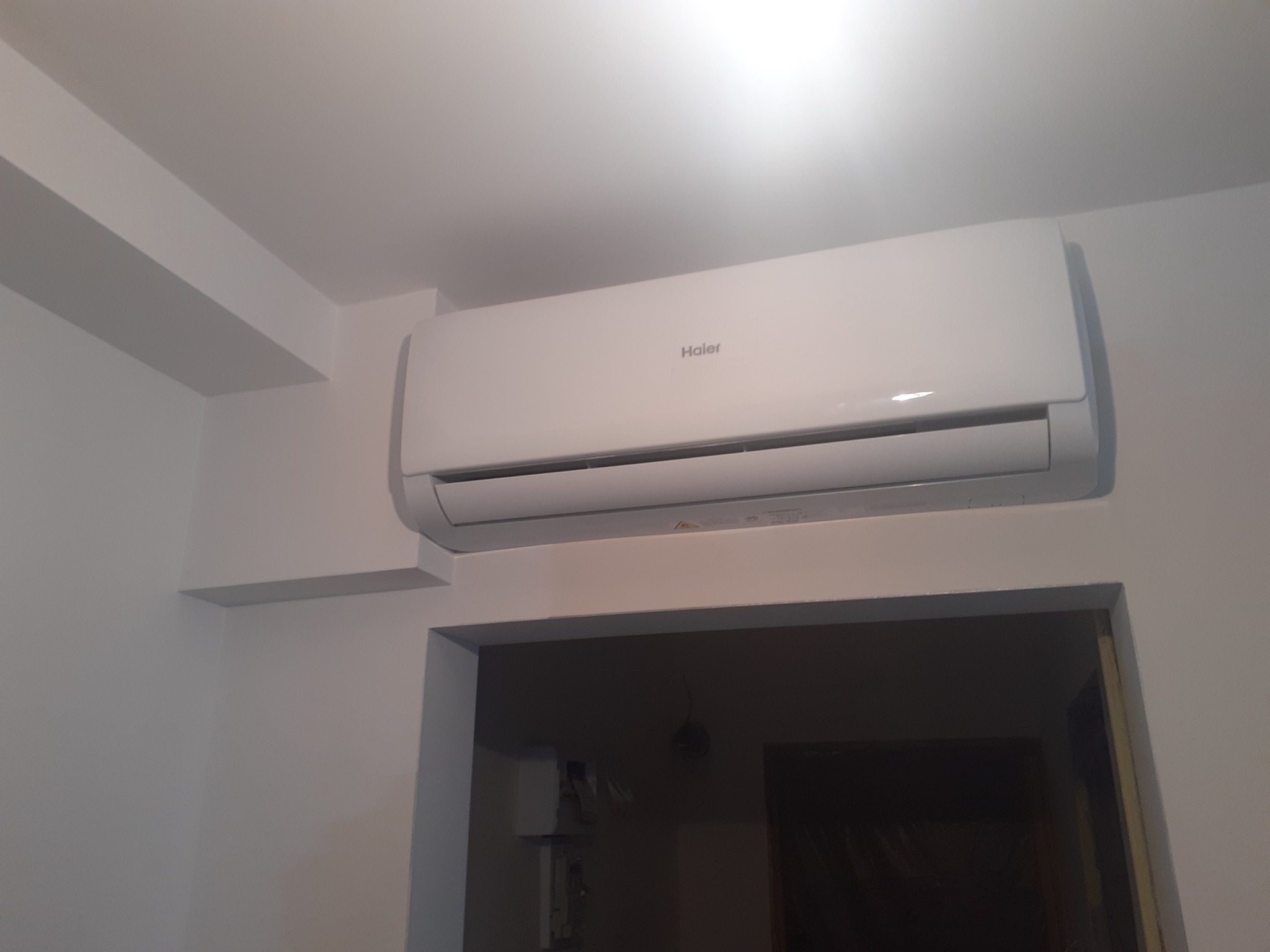 Biała klimatyzacja Haier zamontowana nad drzwiami w jasnym pomieszczeniu, widoczny fragment ściany z półką.