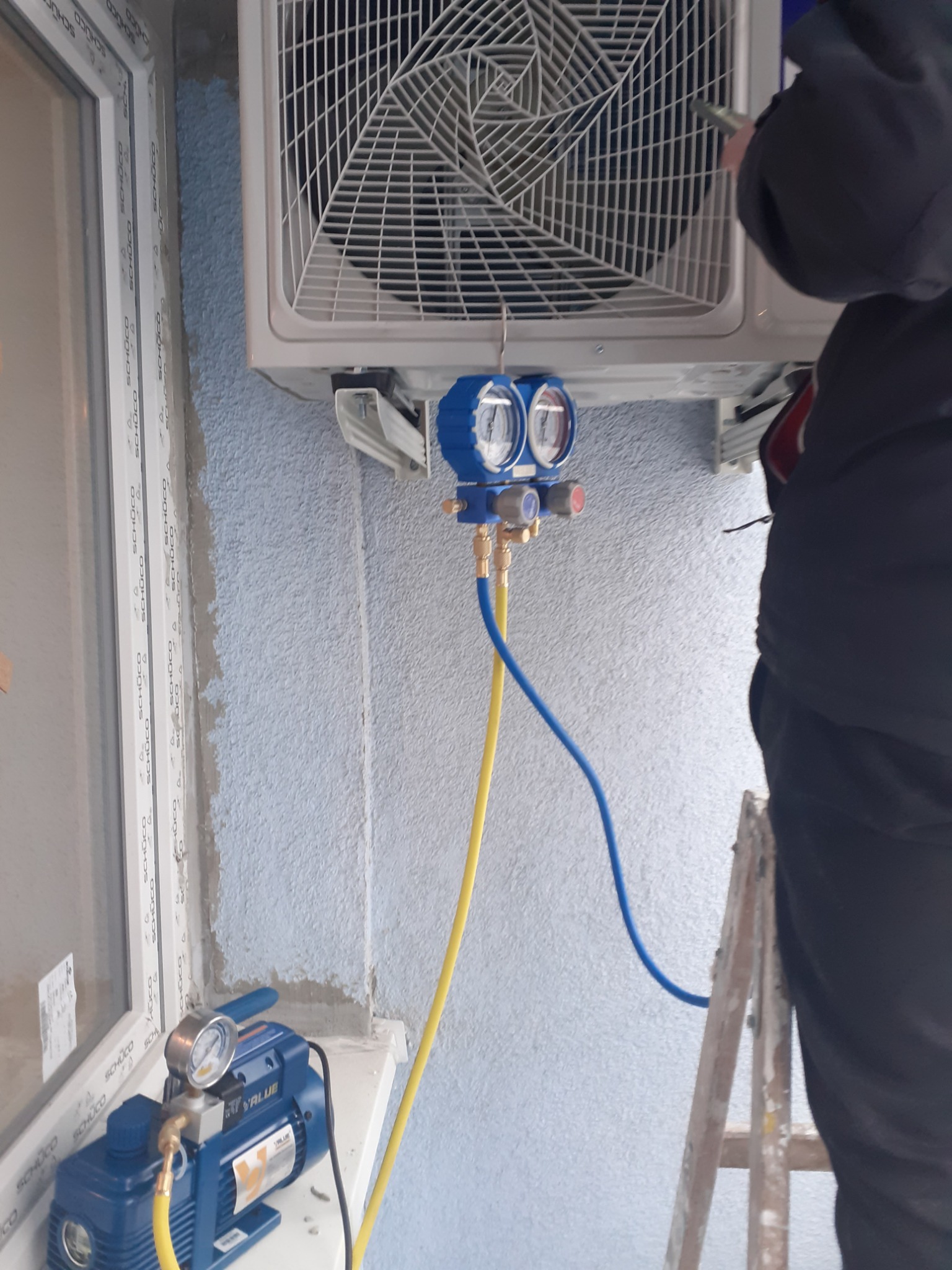 Serwisant na drabinie podłącza manometry do zewnętrznej jednostki klimatyzatora na ścianie budynku, obok okna z ramą SCHUCO. Na parapecie stoi pompa próżniowa VALUE.