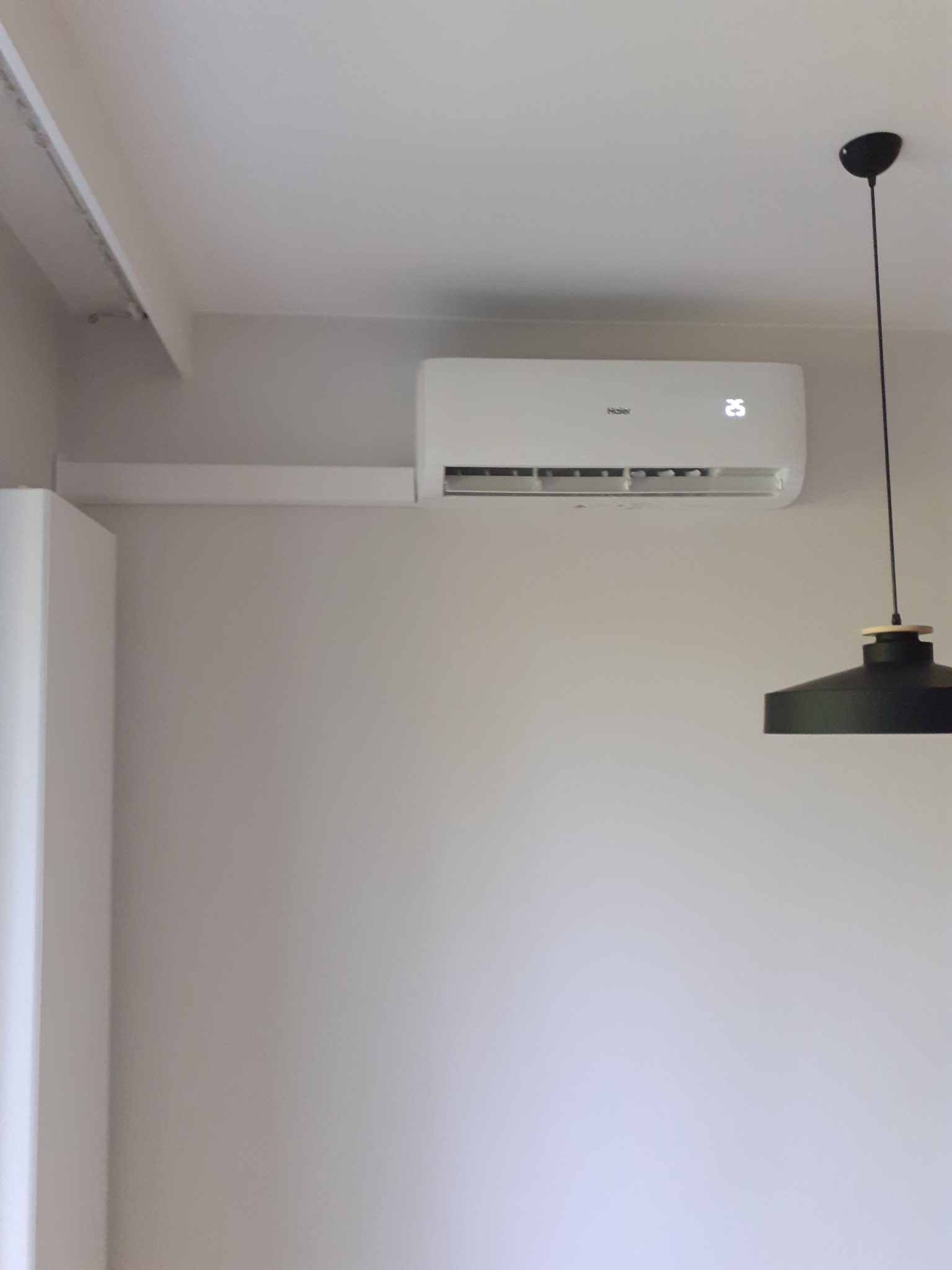 Biały klimatyzator Haier z wyświetlaną temperaturą 25 stopni, zamontowany na ścianie w jasnym pomieszczeniu, widoczna lampa wisząca z czarnym kloszem.