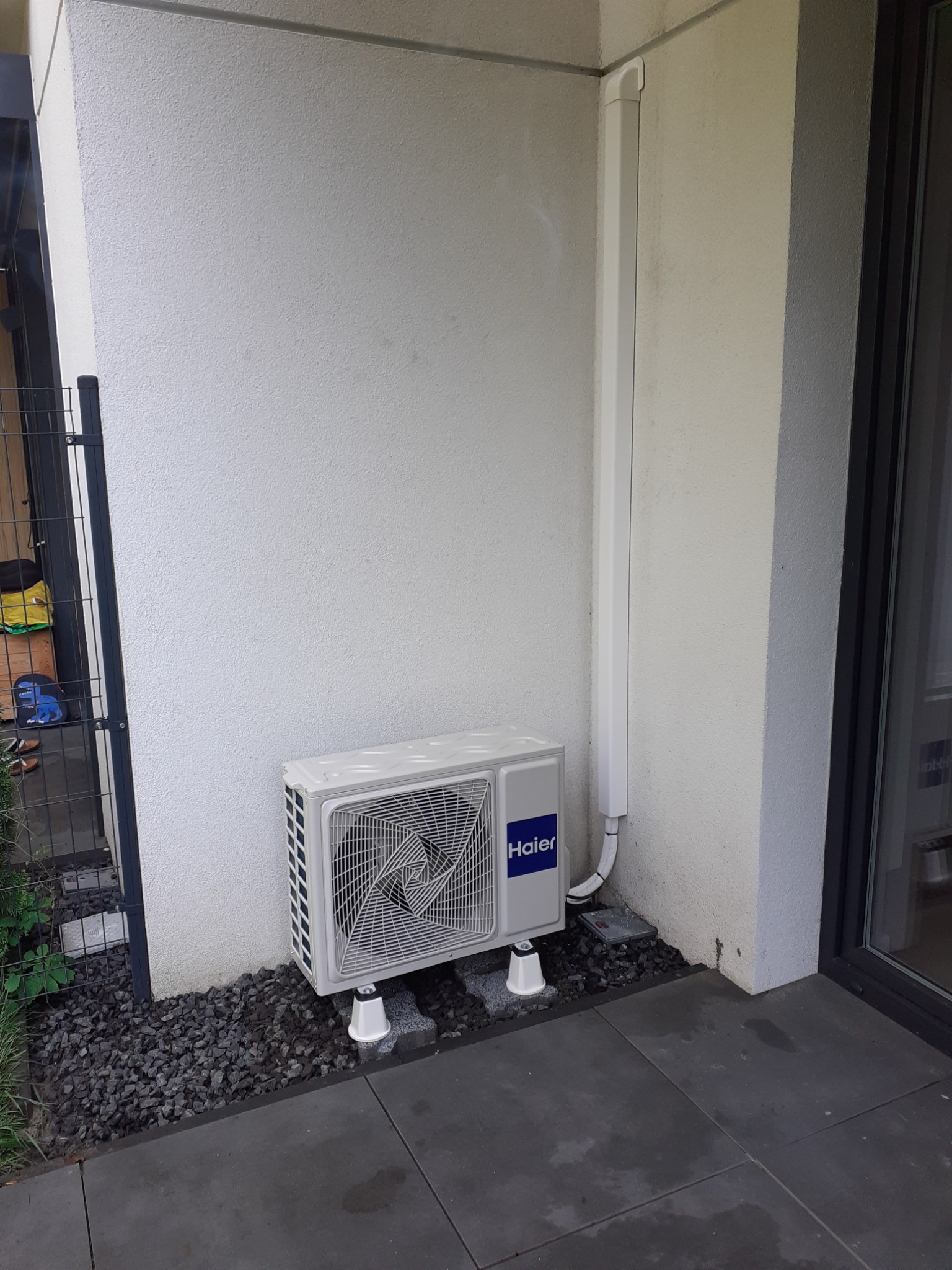 Zewnętrzna jednostka klimatyzatora Haier, zamontowana na białej ścianie domu, z widoczną instalacją odprowadzającą skropliny i ułożonymi kamieniami dekoracyjnymi u podstawy.