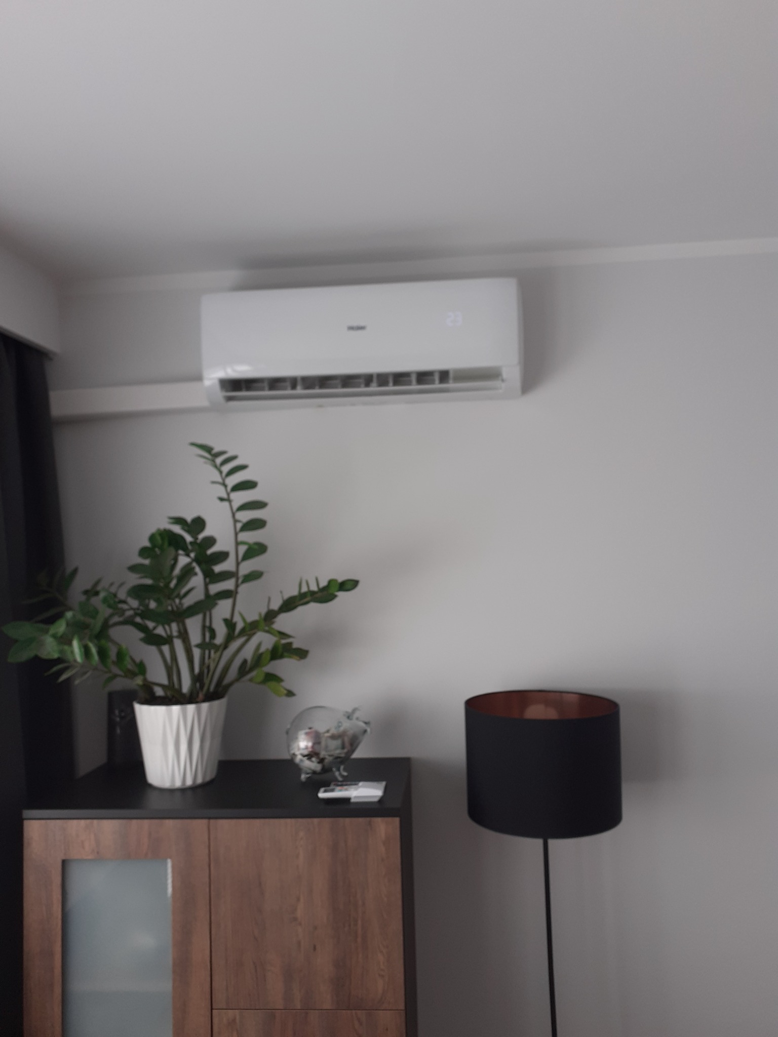 Biały klimatyzator ścienny z wyświetlaną temperaturą 23 stopnie, zamontowany w pokoju nad szafką z rośliną doniczkową i lampą podłogową.