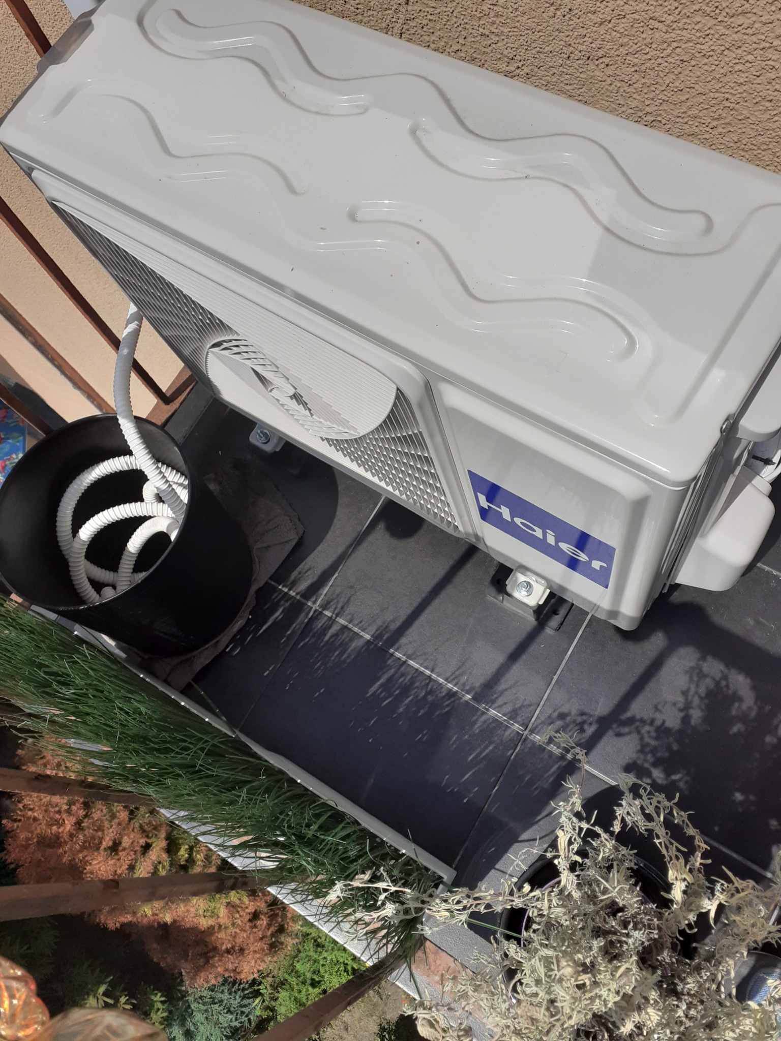 Jednostka zewnętrzna klimatyzatora Haier zamontowana na balkonie, z widocznymi przewodami w czarnym wiadrze i roślinami w tle.