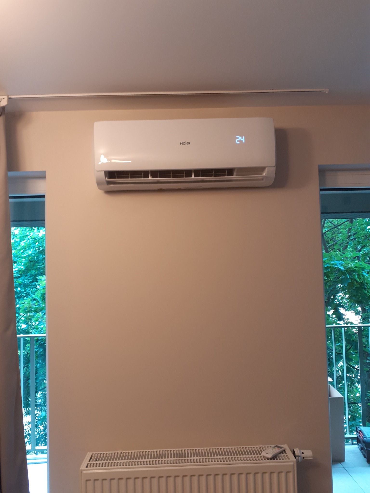 Biała klimatyzacja Haier zamontowana na ścianie w pomieszczeniu, wyświetlająca temperaturę 24 stopnie, widoczny grzejnik pod spodem i zieleń za oknem.