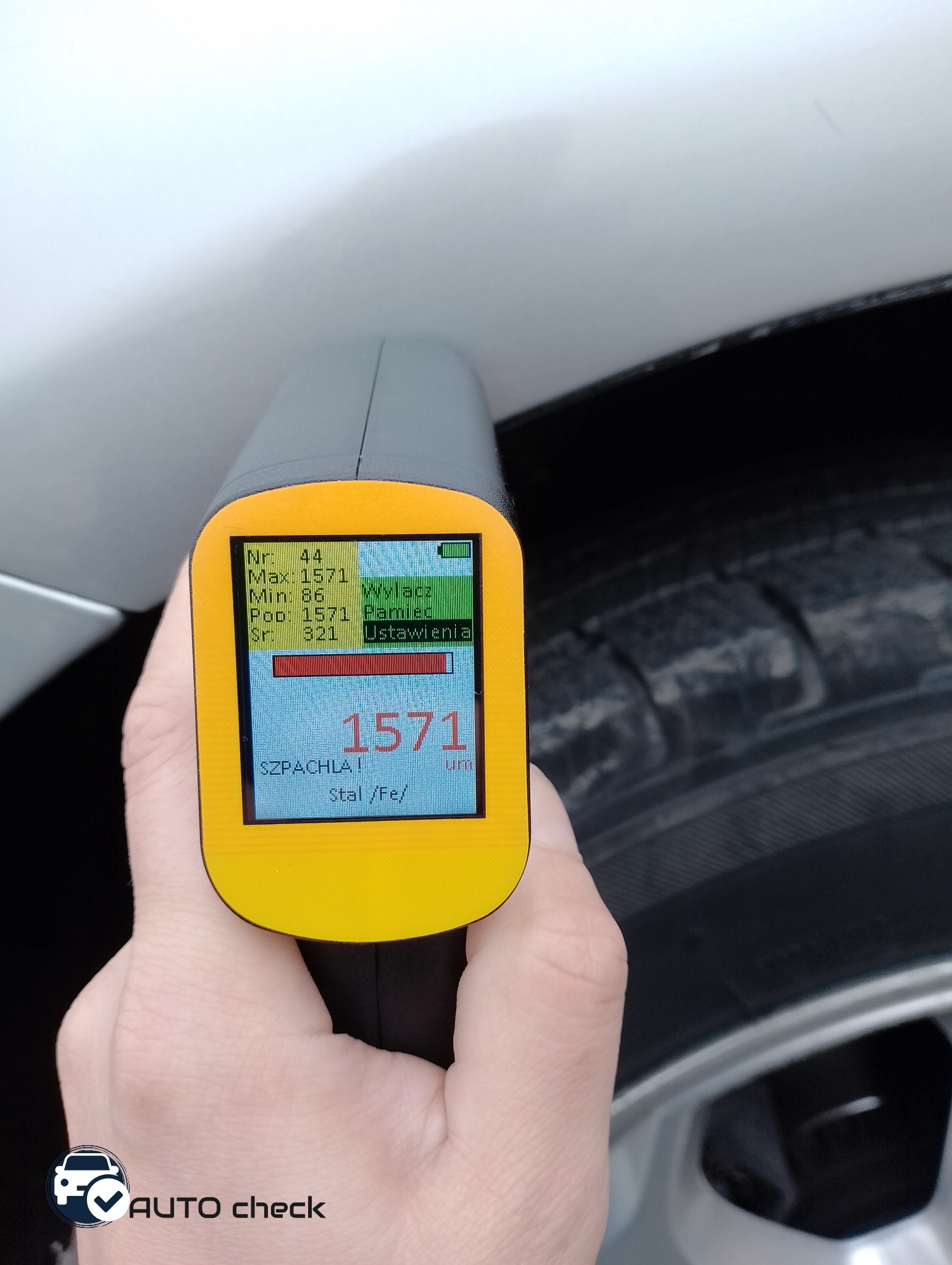Pomiar grubości lakieru na samochodzie za pomocą miernika elektronicznego, wyświetlacz wskazuje wartość 1571 µm, widoczny fragment opony i nadkola.