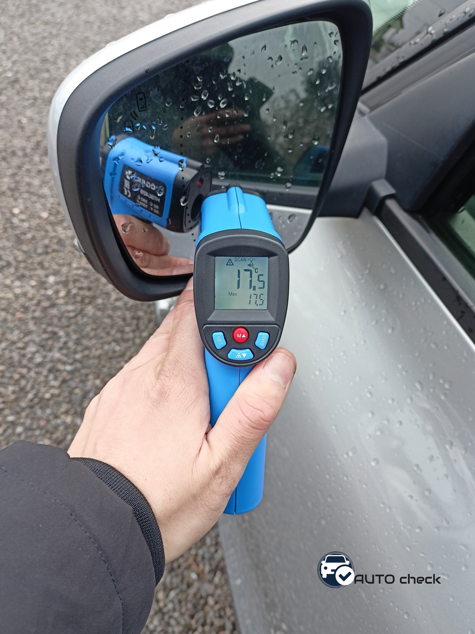 Pomiar temperatury lusterka samochodu za pomocą niebieskiego termometru laserowego, wskazującego 17.5 stopni Celsjusza, widoczne krople deszczu na powierzchni.