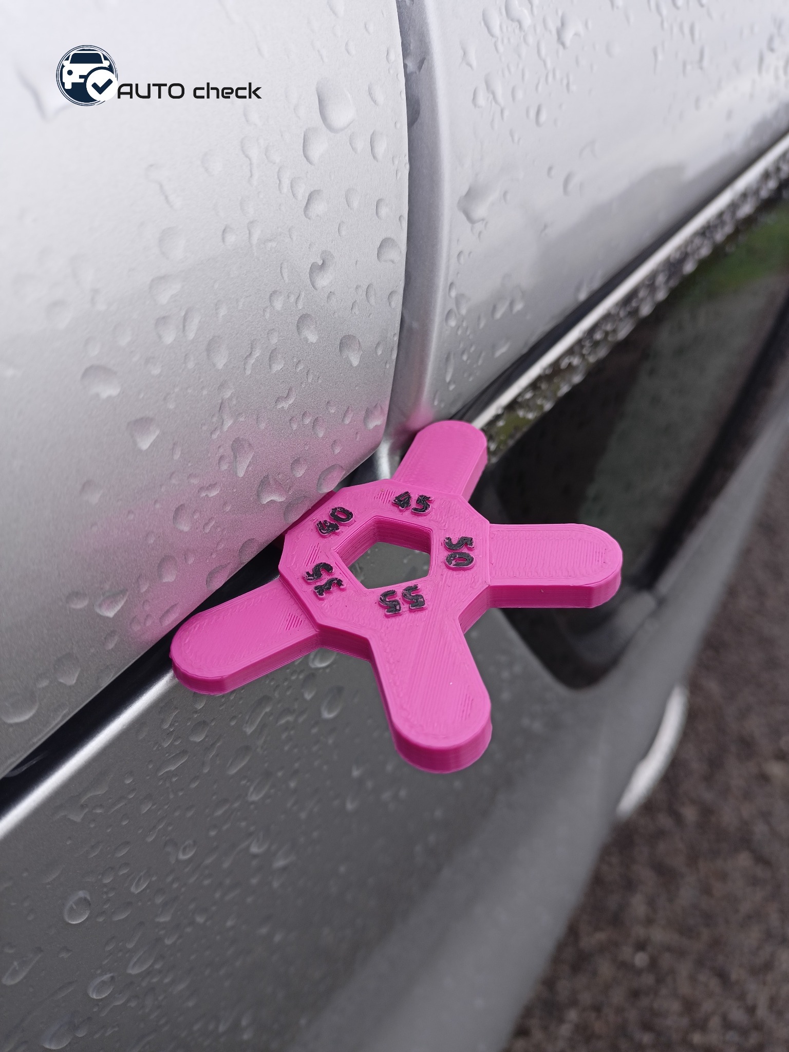 Różowy miernik grubości lakieru z nadrukowanymi wartościami 40, 45, 50, 55, przyłożony do mokrego, srebrnego elementu karoserii samochodu.
