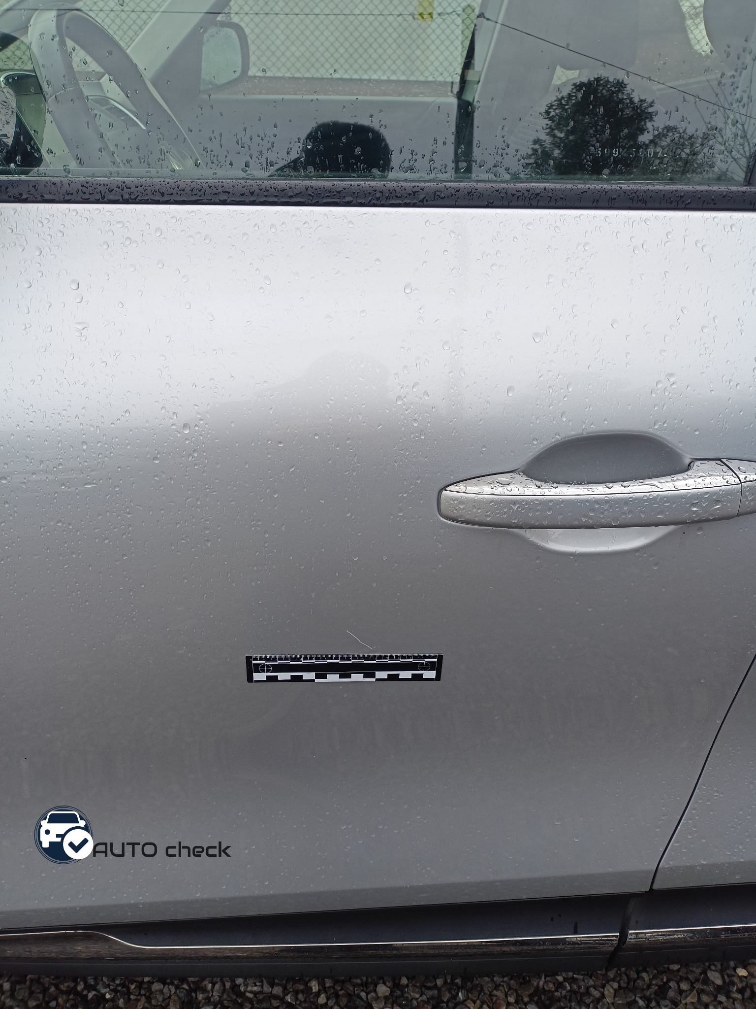 Drzwi samochodu w kolorze srebrnym pokryte kroplami deszczu, z naklejką 'AUTO check' i czarno-białą miarką pomiarową przyklejoną obok.