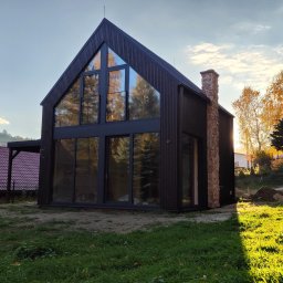 Futurum Sp. Z o.o. - Nowoczesny dom z ciemnego drewna z dużymi oknami i kamiennym kominem w jesiennym słońcu.
