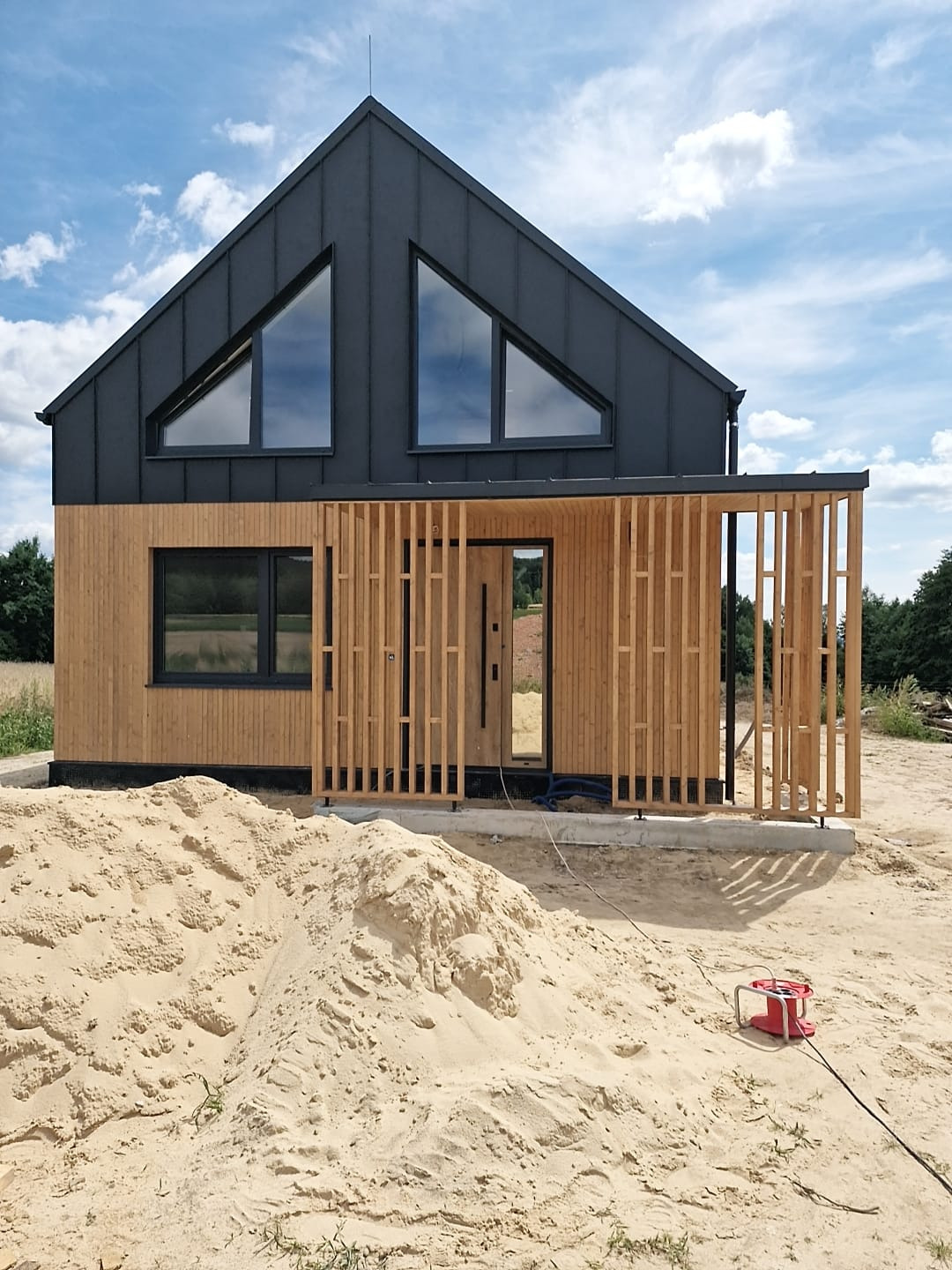 Nowoczesny dom z ciemnym, spadzistym dachem i drewnianą elewacją, częściowo osłonięty drewnianą konstrukcją; na pierwszym planie hałda piasku i czerwony przedłużacz.