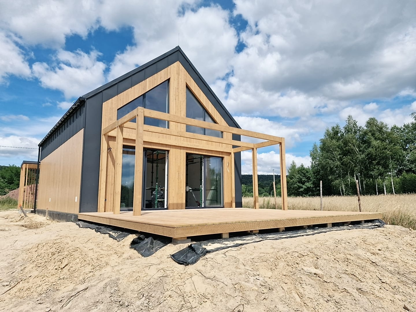 Nowoczesny dom z drewnianą elewacją i dużym tarasem, widok z poziomu gruntu na tle błękitnego nieba z chmurami.