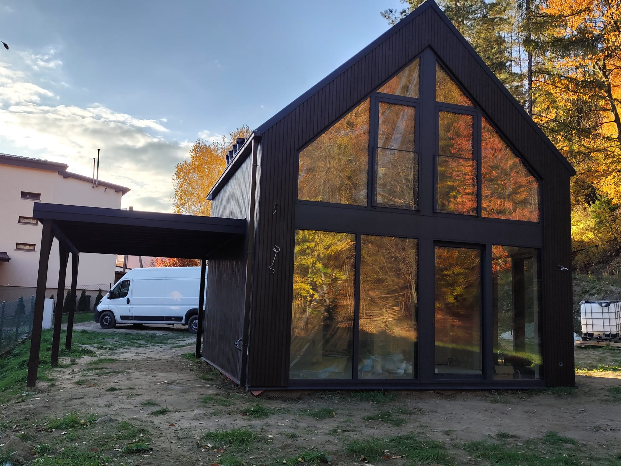 Nowoczesny, ciemnobrązowy dom z dużymi oknami, w których odbijają się jesienne drzewa, z dobudowaną wiatą i białym samochodem dostawczym w tle.