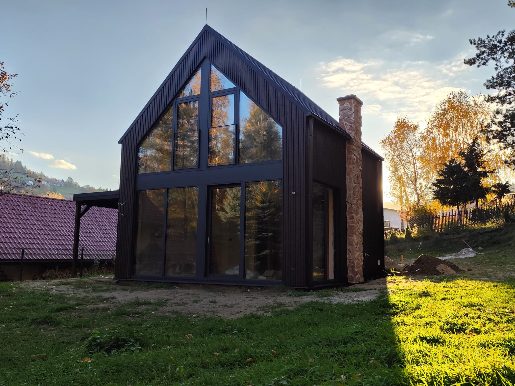 Nowoczesny dom z ciemnego drewna z dużymi oknami i kamiennym kominem w jesiennym słońcu.