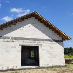 SBC Adam Bączkowski - Wznoszenie ścian parterowego domu z bloczków, widoczne drewniane elementy konstrukcji dachu i nieotynkowane ściany, w tle błękitne niebo i las.