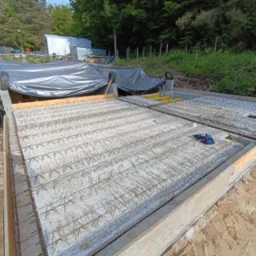 Zbrojona płyta fundamentowa w szalunku, widok z góry. W tle zieleń i budynki gospodarcze pod plandeką. Prace ziemne w toku, widoczny piasek i ziemia.