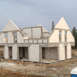 SBC Adam Bączkowski - Surowy stan domu z białego betonu komórkowego, widoczne otwory okienne i drzwiowe, betonowe nadproża, teren budowy z gruzem i materiałami budowlanymi.
