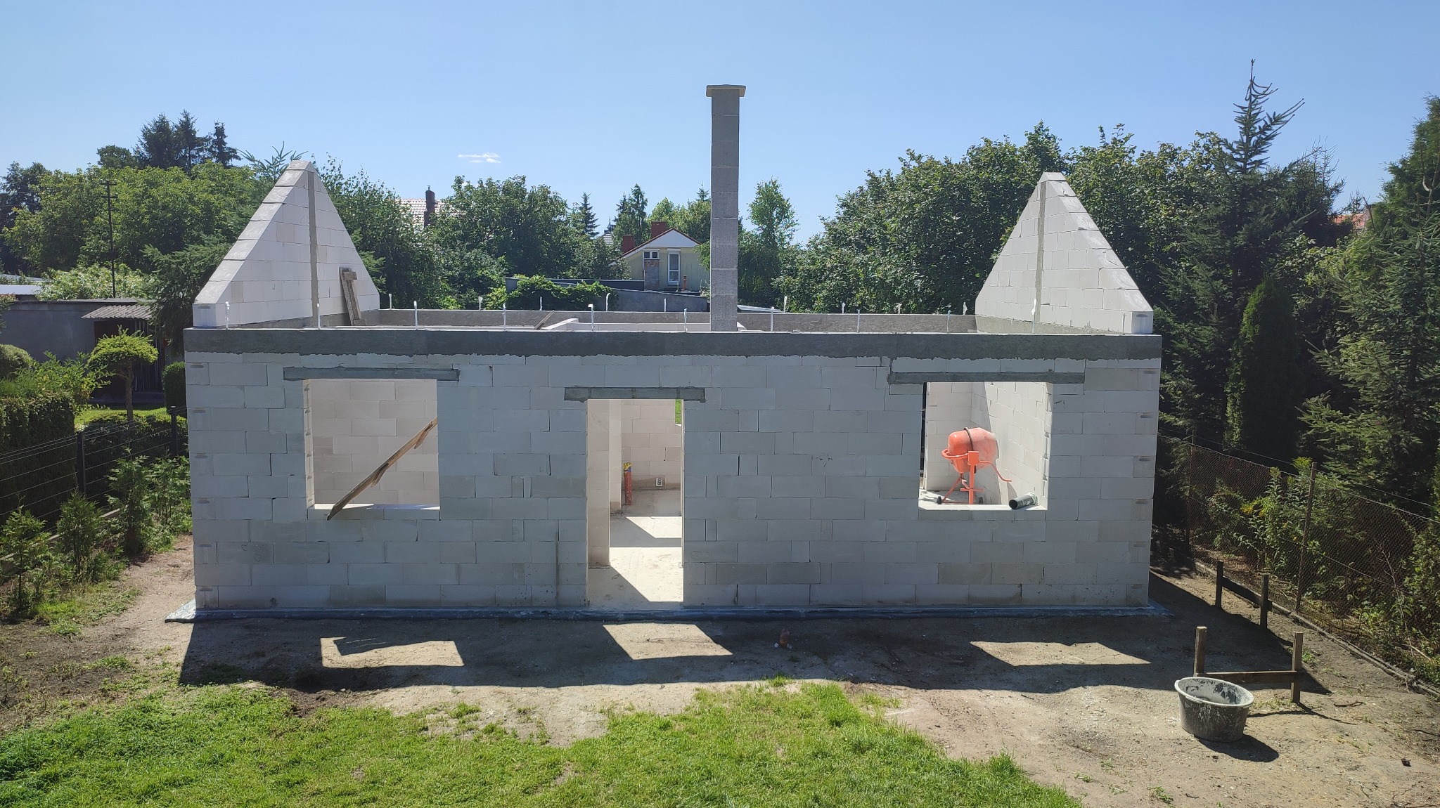 Wznoszenie ścian parterowego domu z betonu komórkowego, widoczny komin, przygotowane otwory okienne, w jednym z nich stoi betoniarka. Na zewnątrz zieleń i fragment zabudowy.