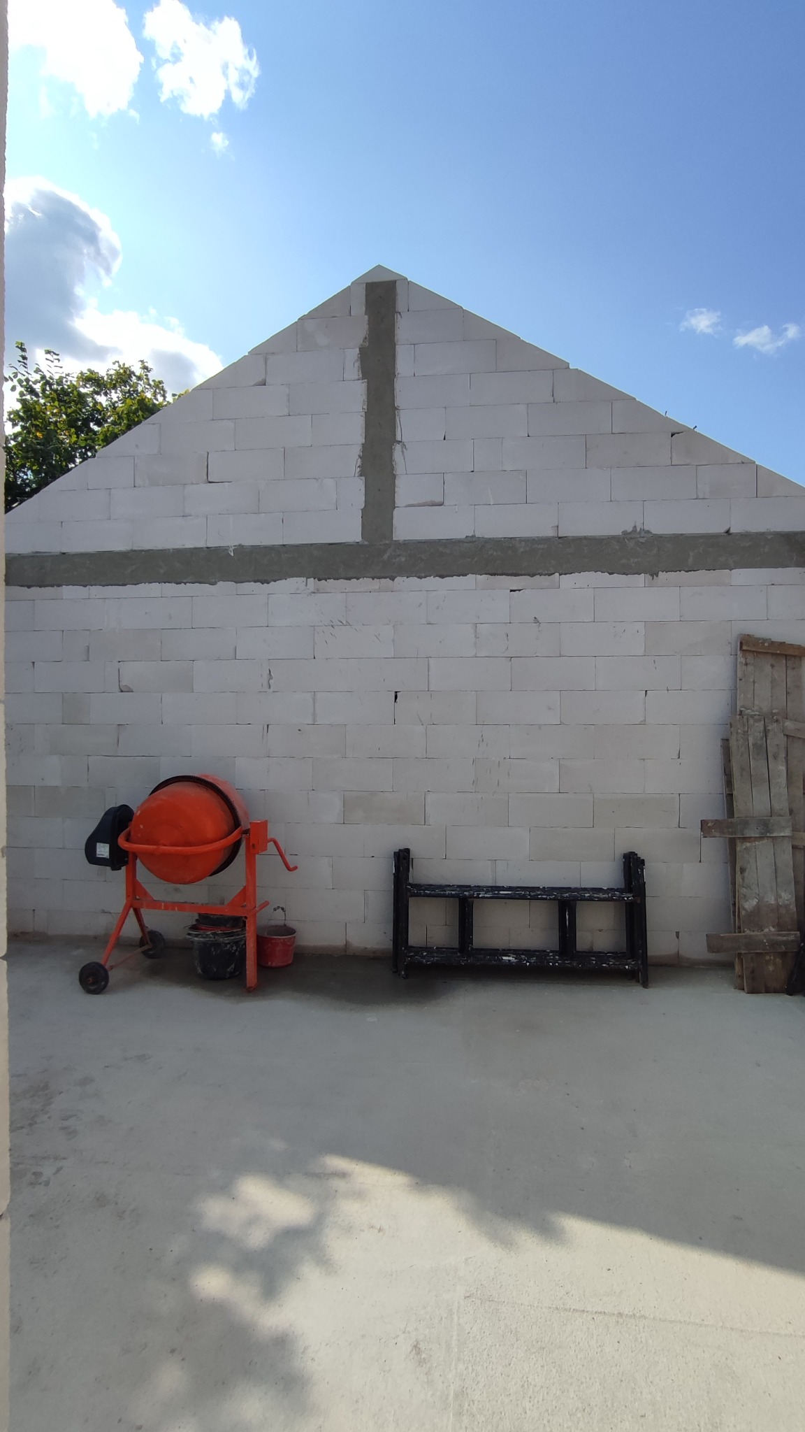 Wznoszenie ścian domu z bloczków, widoczna pomarańczowa betoniarka, czarny stojak i deski oparta o ścianę. Na ścianie pionowe i poziome wzmocnienia z zaprawy.