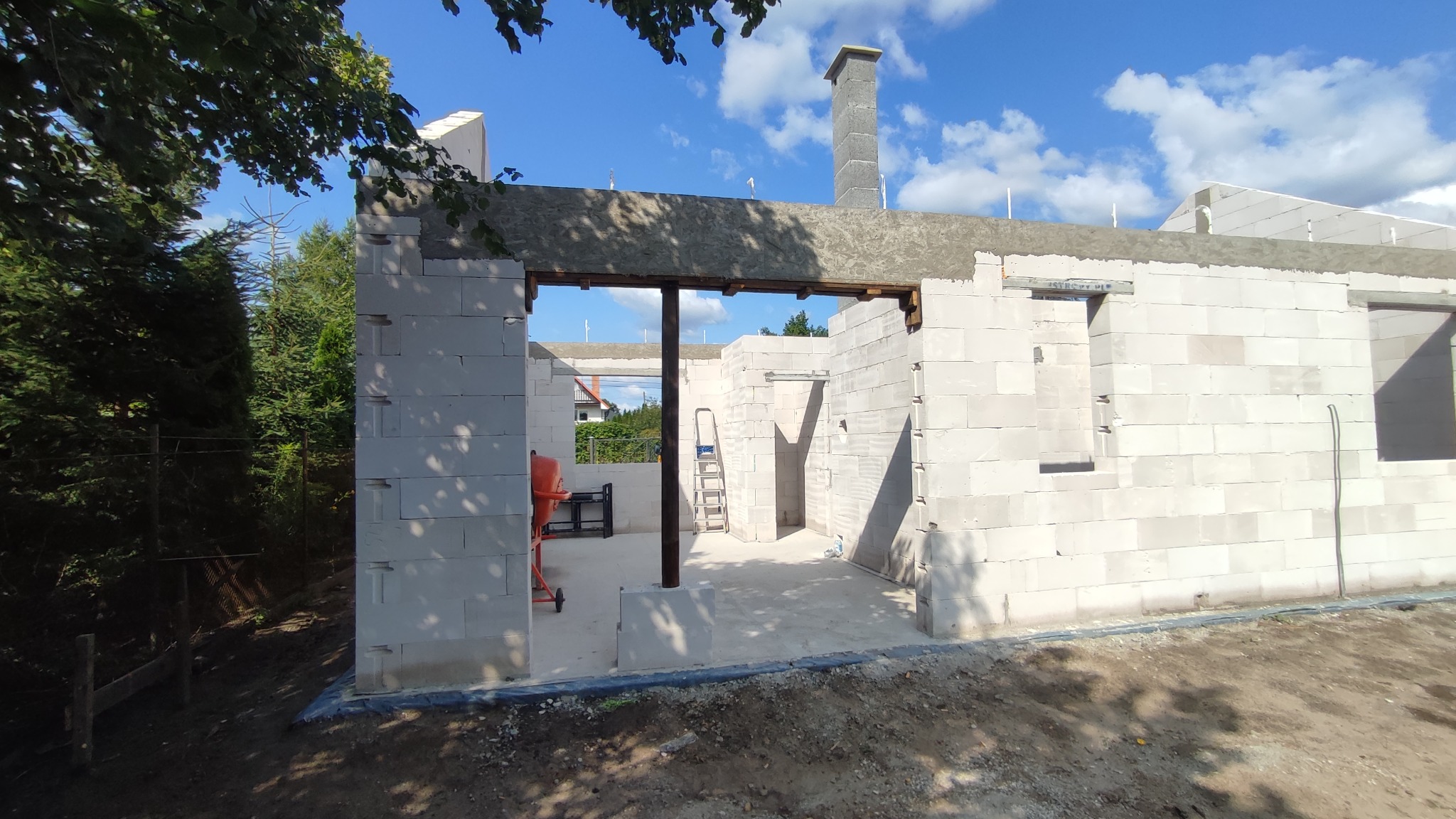 Wznoszenie ścian domu z białych bloczków, widoczny komin, mieszalnik do betonu i drabina w surowym wnętrzu, niebo z chmurami w tle.