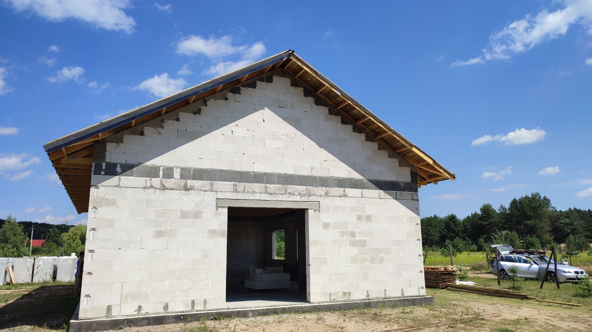 Wznoszenie ścian parterowego domu z bloczków, widoczne drewniane elementy konstrukcji dachu i nieotynkowane ściany, w tle błękitne niebo i las.