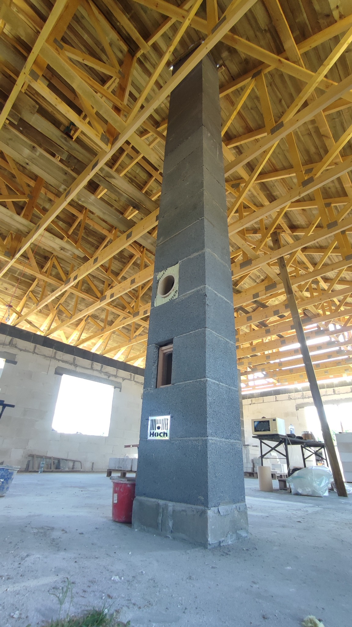 Wznoszący się w górę komin z szarej bloczków w surowym stanie, z widocznymi otworami i logo producenta, na tle drewnianej konstrukcji dachu w trakcie budowy.