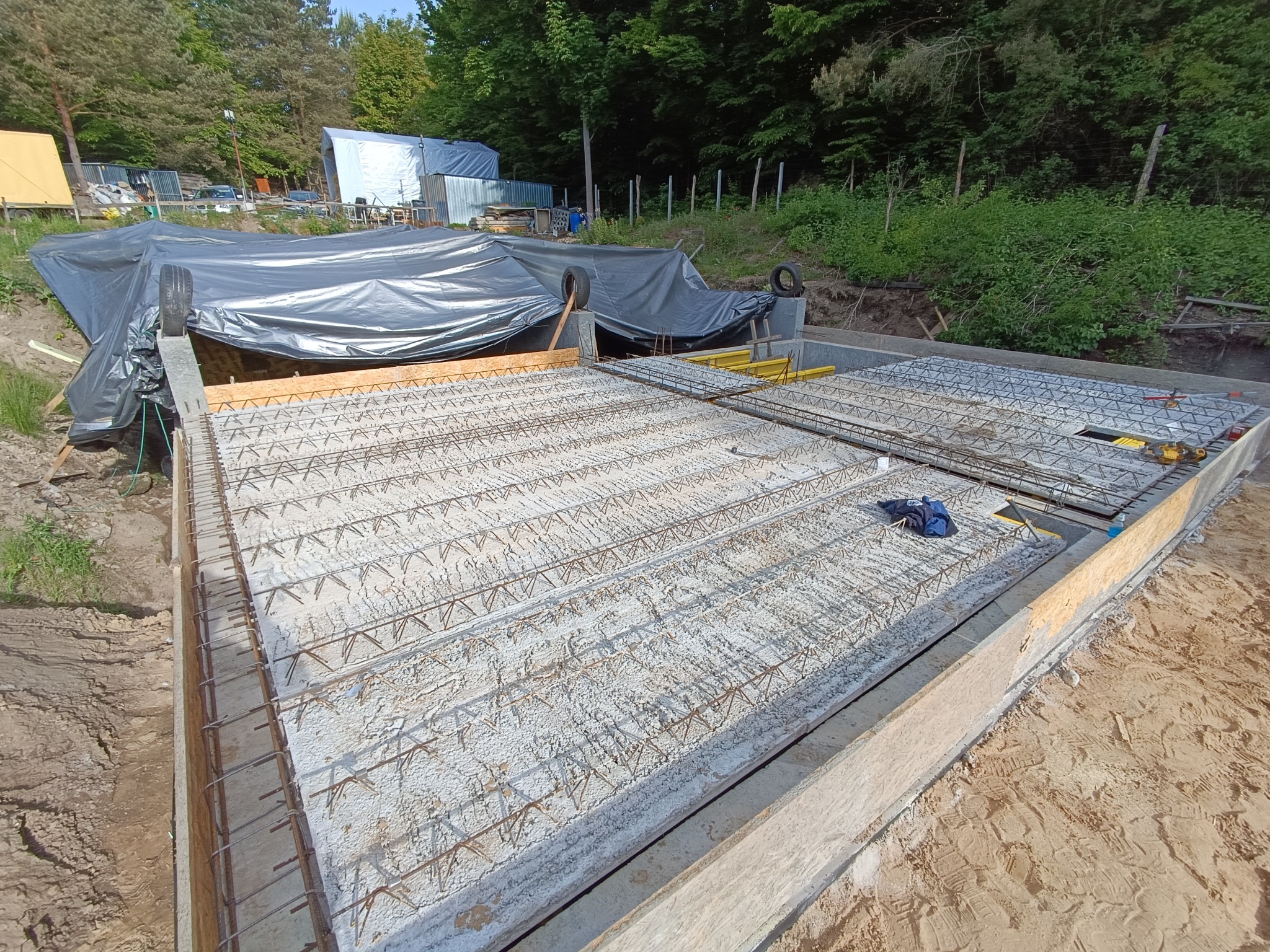 Zbrojona płyta fundamentowa w szalunku, widok z góry. W tle zieleń i budynki gospodarcze pod plandeką. Prace ziemne w toku, widoczny piasek i ziemia.