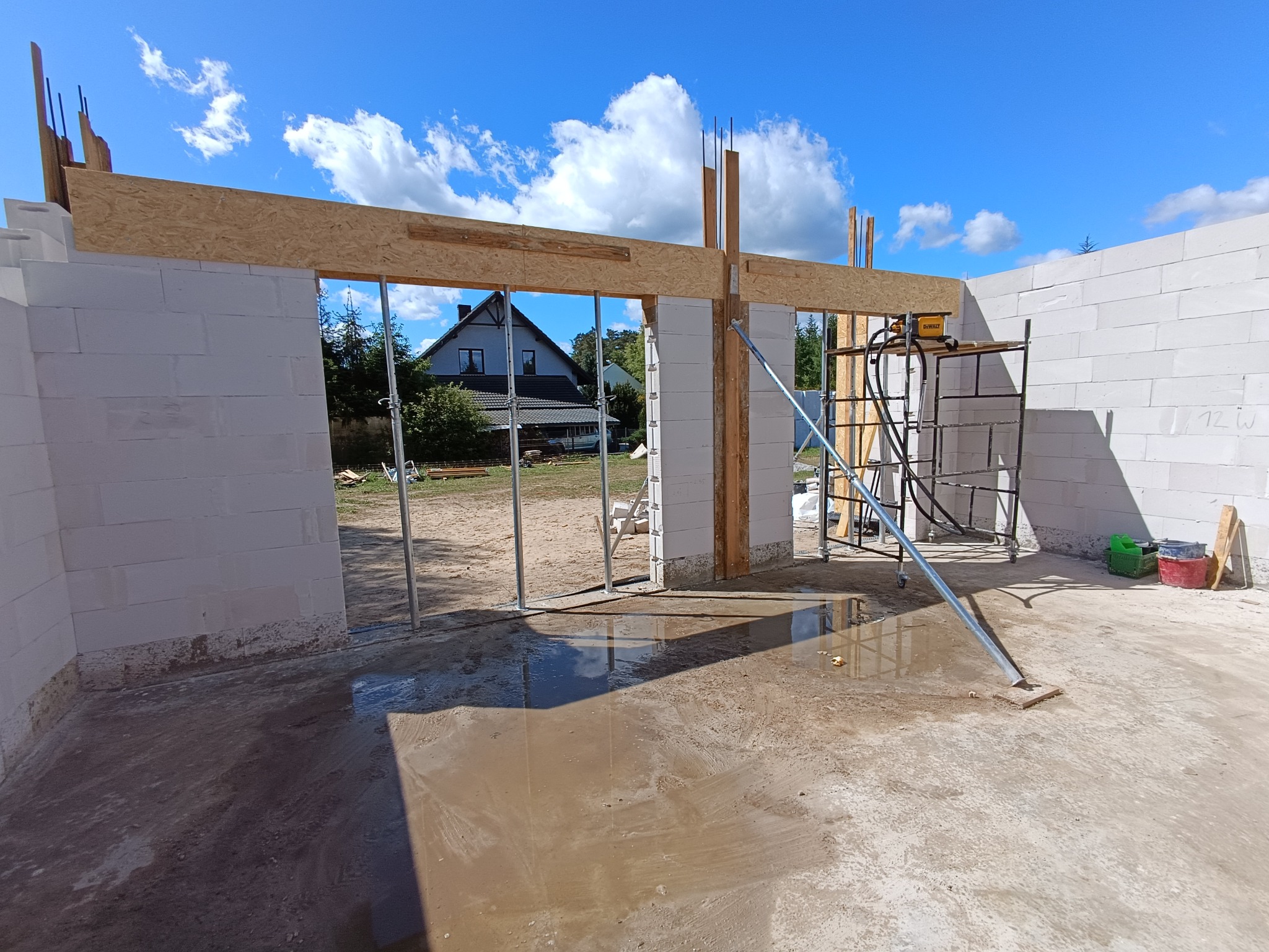 Budowa domu z białych bloczków: widok na ściany z otworami okiennymi i drzwiowymi, wsparte drewnianą konstrukcją. W tle dom jednorodzinny i błękitne niebo.