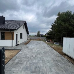KM-BUD Marcel Kmieć - Długi podjazd z szarych płyt betonowych o nieregularnym wzorze, prowadzący do nowoczesnego, białego domu z czarnym dachem. Widoczne świeżo ułożone płyty i piasek wokół.
