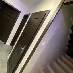 KM DESIGN Kamil Mering - Ciemne, drewniane drzwi wewnętrzne z srebrną klamką, zamontowane w jasnym pomieszczeniu z widocznymi schodami i fakturą na ścianie.