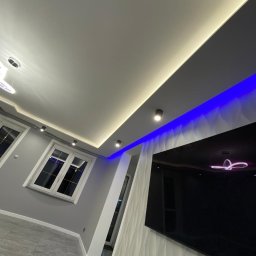 KM DESIGN Kamil Mering - Nowoczesne wnętrze z podwieszanym sufitem z oświetleniem LED (białym i niebieskim), szarą ścianą, białymi oknami i telewizorem na ozdobnej ścianie.