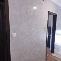 Ściana w korytarzu z dekoracyjną, szarą strukturą, widoczny włącznik światła, czarna futryna drzwi wejściowych i futryna drzwi do pomieszczenia z widocznym panelem sterowania.