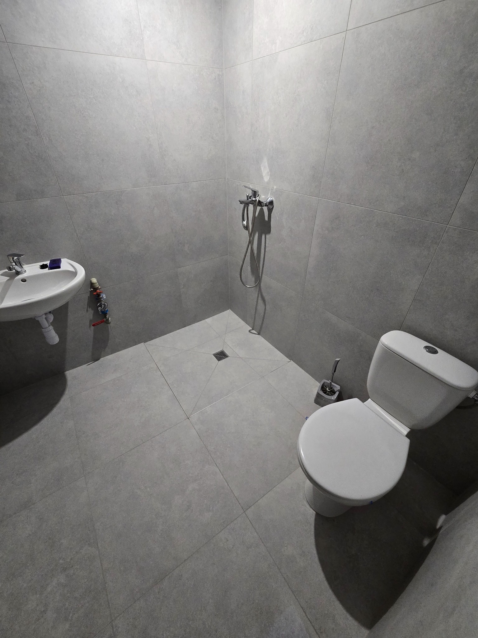 Szara łazienka z białą ceramiką: umywalka, toaleta, bateria prysznicowa, płytki wielkoformatowe na ścianach i podłodze, odpływ liniowy w rogu kabiny.