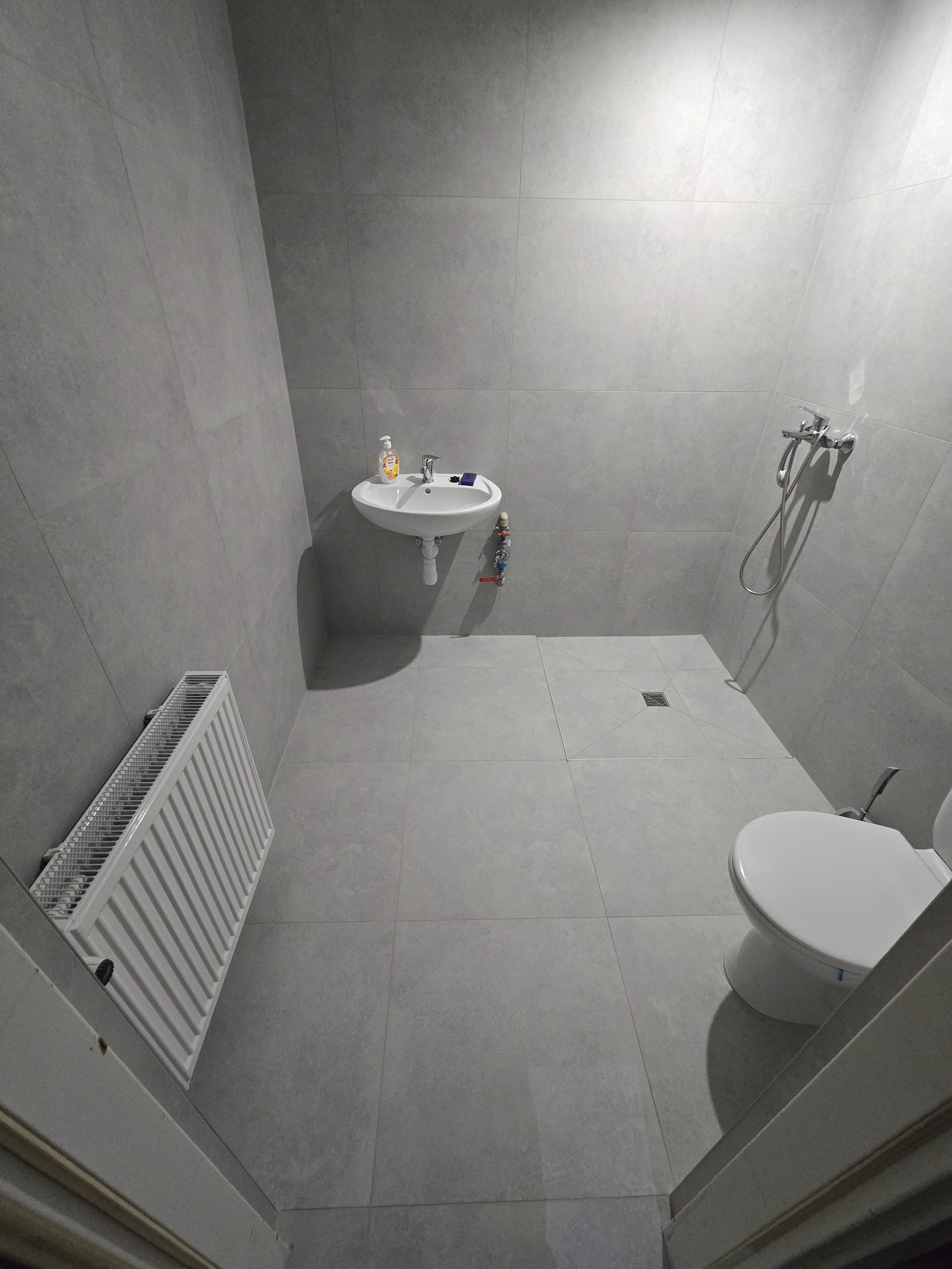 Nowoczesna łazienka wykończona w szarościach, z umywalką, grzejnikiem i toaletą. Widoczny odpływ liniowy w podłodze i minimalistyczna bateria prysznicowa.