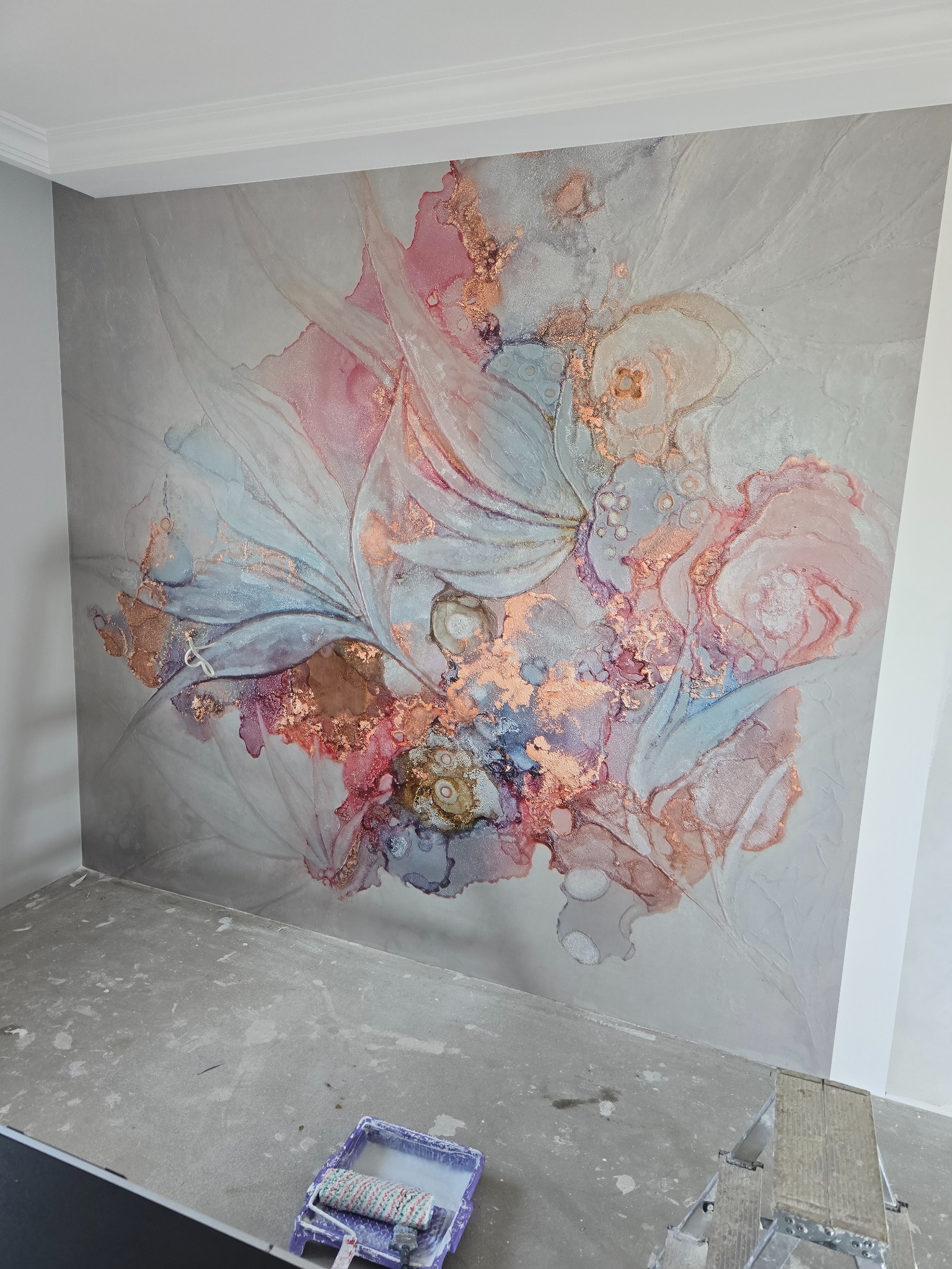 Ściana z dekoracyjną, abstrakcyjną tapetą w odcieniach różu i szarości z elementami złota, wykonana w Borowie, widoczny wałek malarski i drabina, sugerujące świeżo zakończone wykończenie.