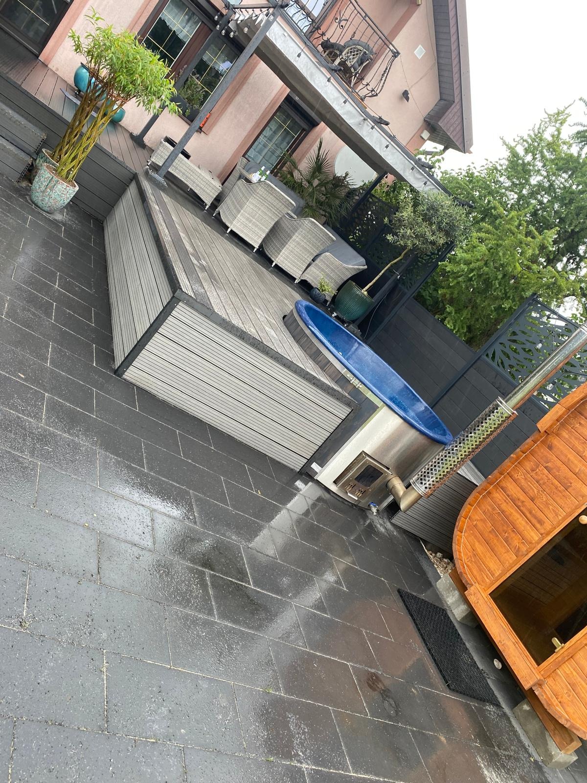 Nowoczesna aranżacja tarasu z drewnianą balią z piecem opalanym drewnem, szarymi płytkami i meblami ogrodowymi z technorattanu, uzupełniona zielenią w donicach.