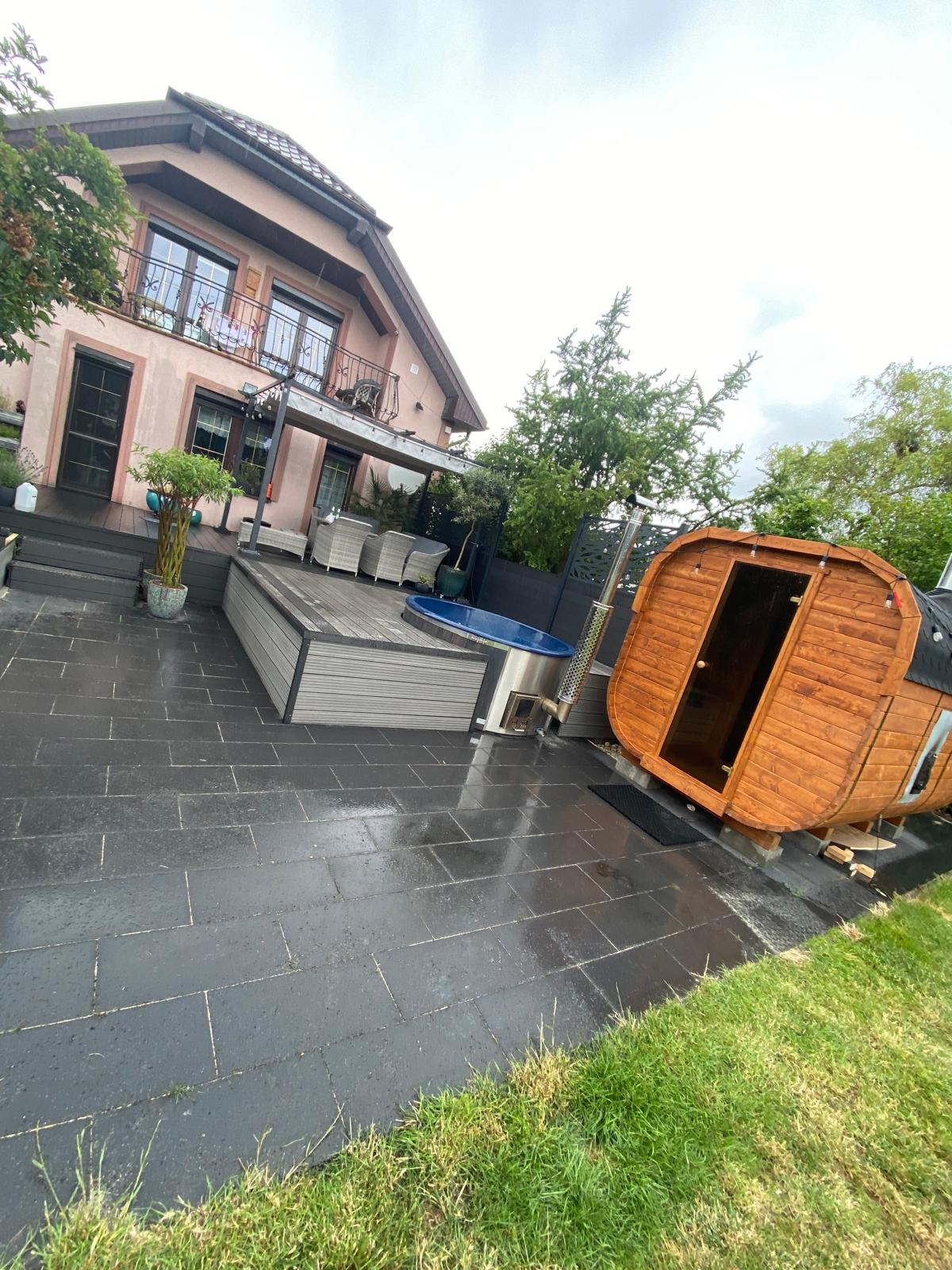 Nowoczesny ogród z drewnianą sauną beczką, jacuzzi i tarasem z szarej kostki brukowej na tle domu z balkonami.