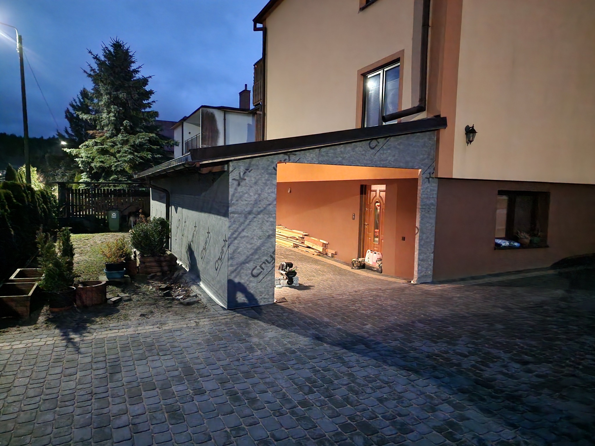 Nowo wybudowana wiata garażowa z widoczną warstwą izolacyjną na ścianach zewnętrznych, wewnątrz ułożone deski i sprzęt budowlany, kostka brukowa na podjeździe