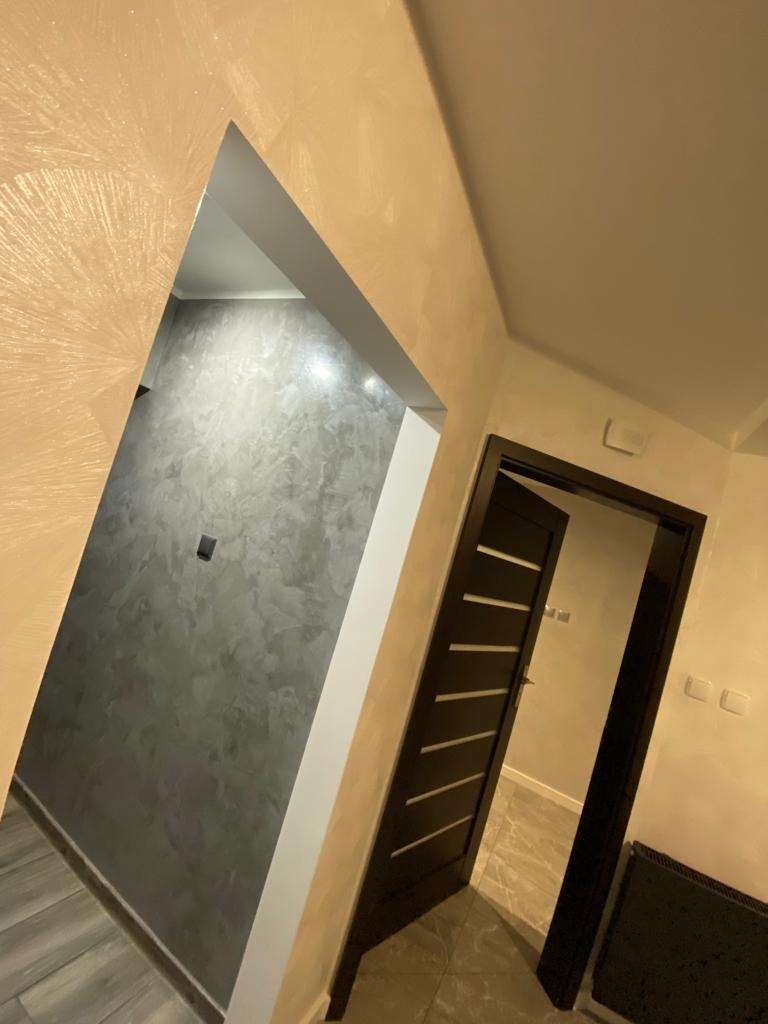 Nowoczesne wnętrze z otwartymi drzwiami, ściany pokryte dekoracyjną, beżowo-złotą fakturą, widoczna szara ściana w pomieszczeniu obok z zamontowanym gniazdkiem elektrycznym, ciemne drzwi...